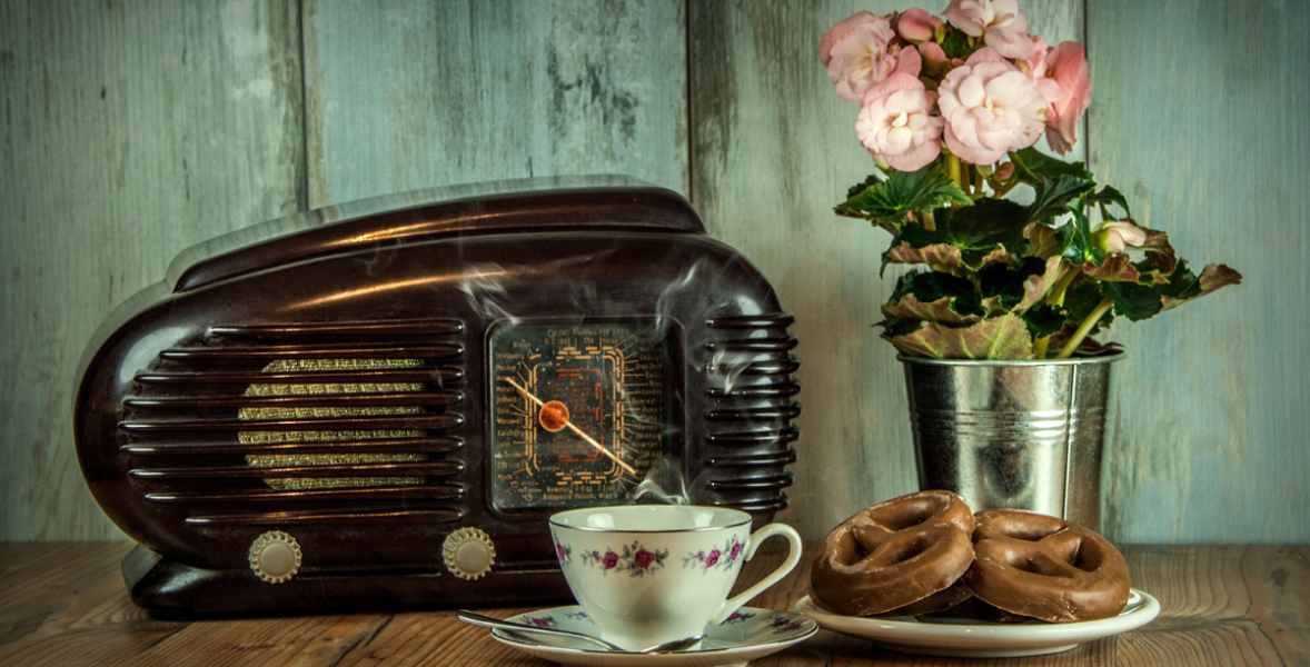 Image Description: vintage looking photograph of an old black dial-radio on a wood table. Next to the radio is a vintage floral china teacup, plate of soft pretzals, and a metal planter with pink peonies growing. Behind the table is a wood wall stained turquoise.